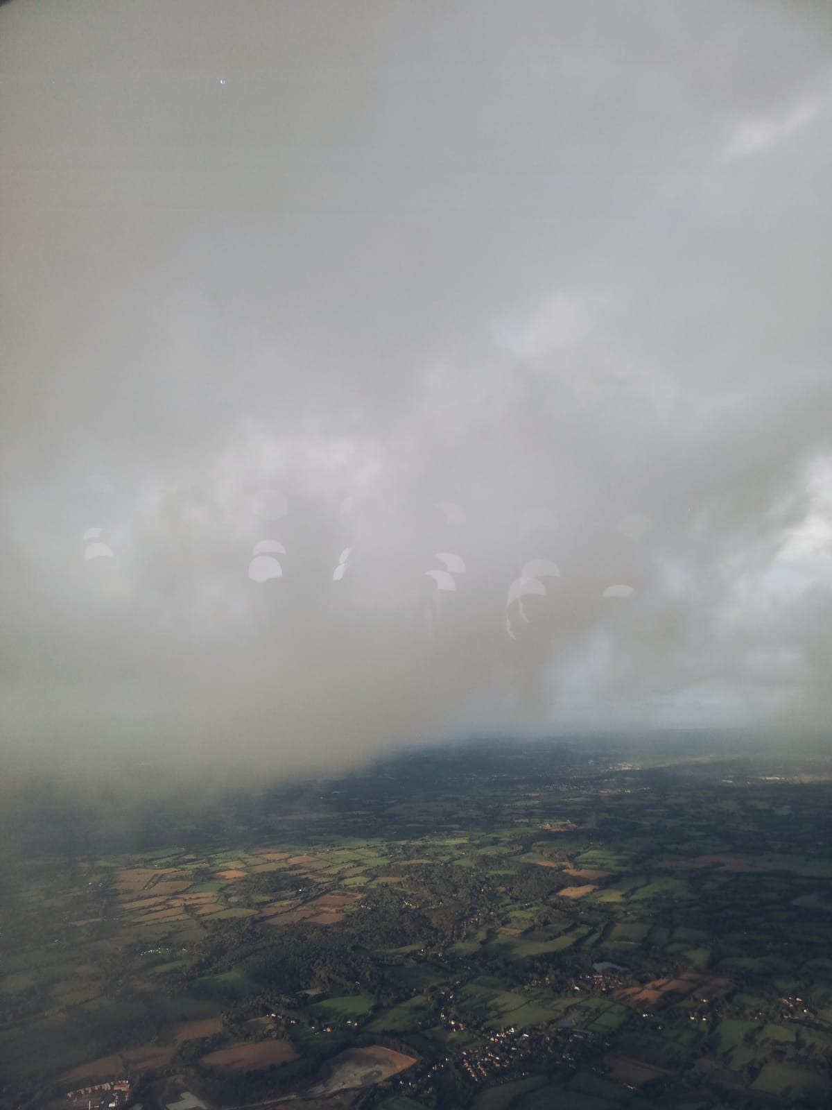 foto tomada desde la ventana de un avión donde se ven prácticamente solo nubes espesas y grises y un terreno abajo mayoritariamente de un verde bosque
