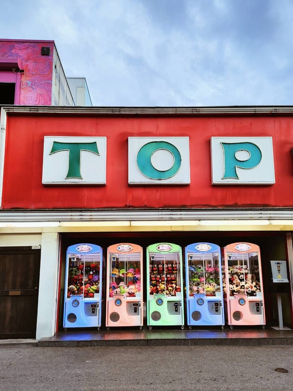 un edificio rojo con letras de neón apagadas que dicen TOP, cubre cinco máquinas de peluches de feria de colores azul, naranja y verde neón